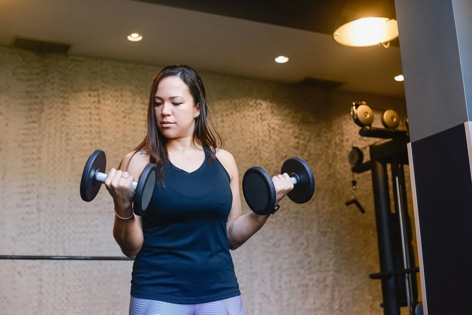 A woman in a black tank top curling a pair of dumbbells, elbows at her sides, in a dimly lit gym.