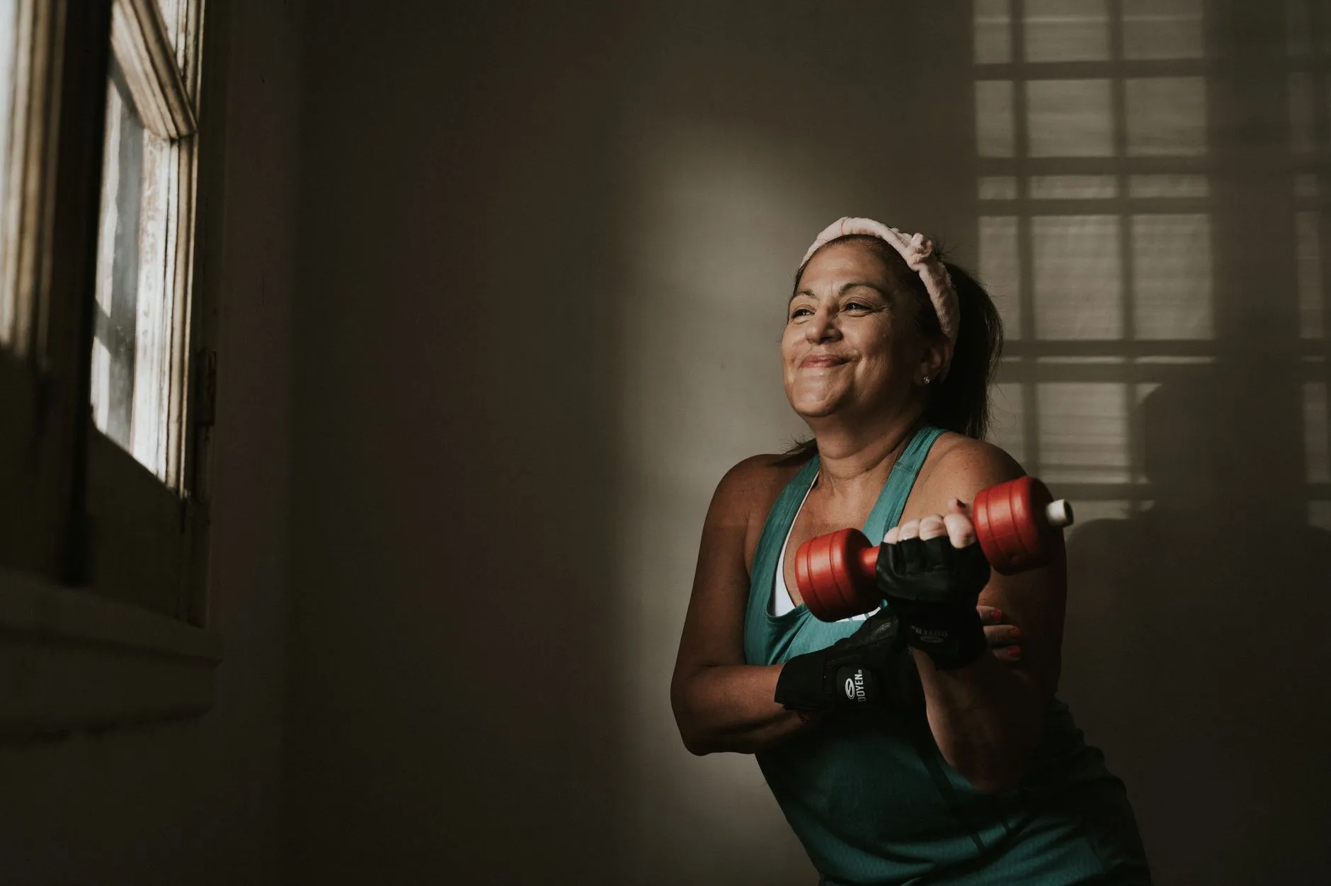 A smiling middle-aged woman in a green tank top lifts a small red dumbbell in a sunlit room at home.