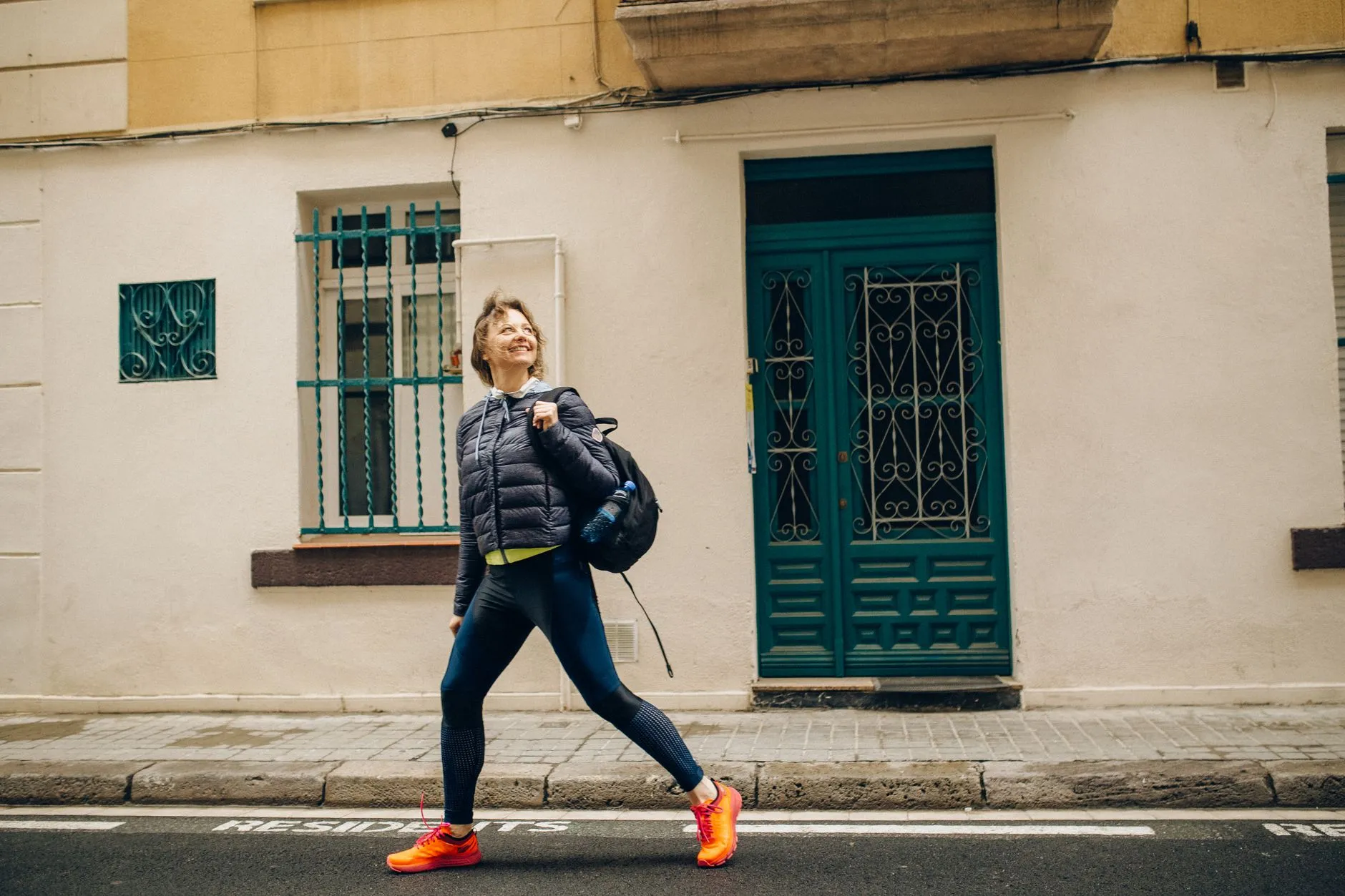 A woman in a jacket and running shoes walking briskly down a city street, smiling and looking off to the side.