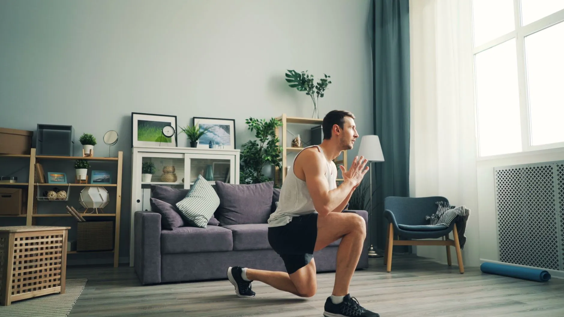 A man in a living room mid-lunge, front knee bent, looking ahead past a sofa and window.
