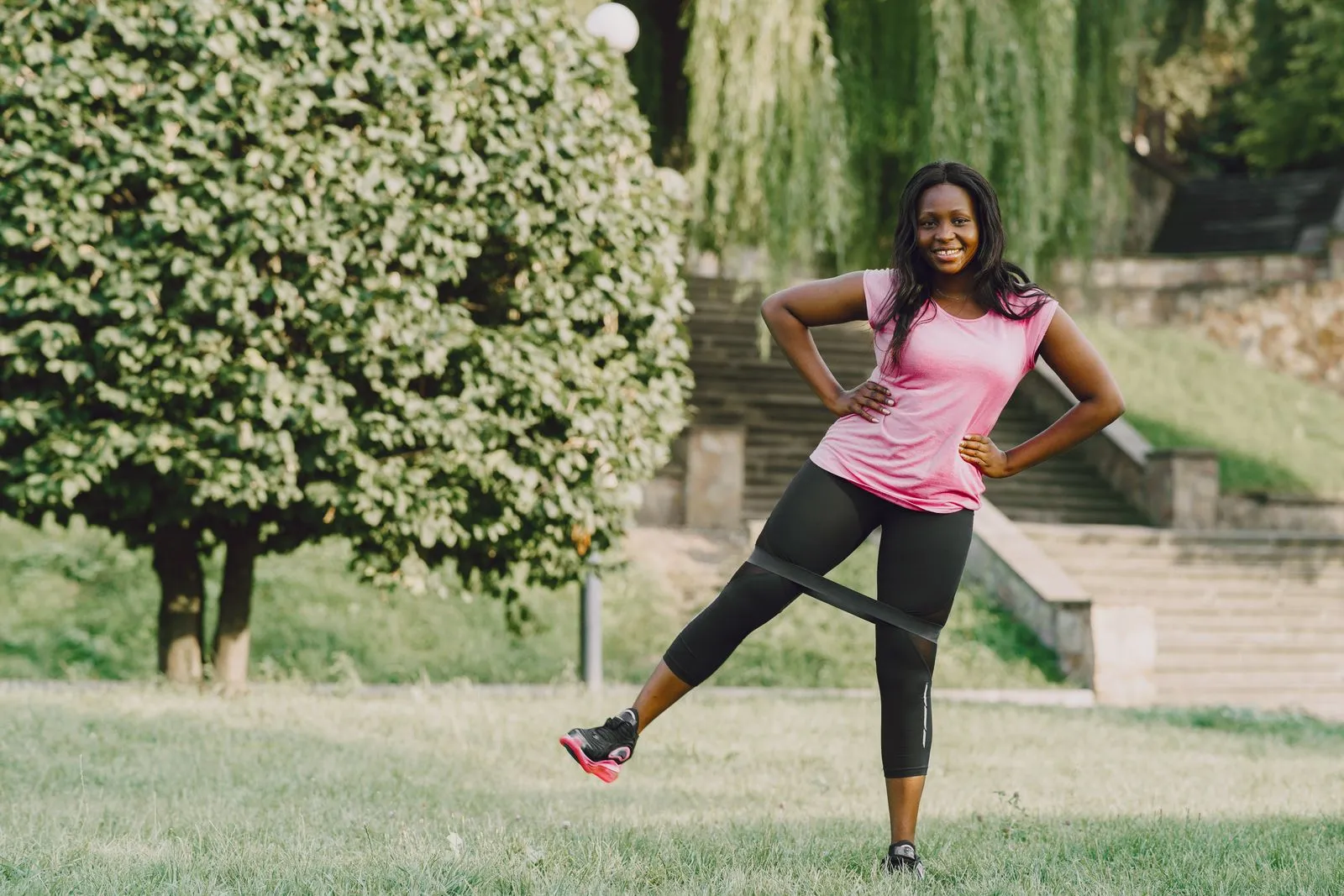 A smiling woman in a sunny park doing a leg exercise with a resistance band looped around her legs.
