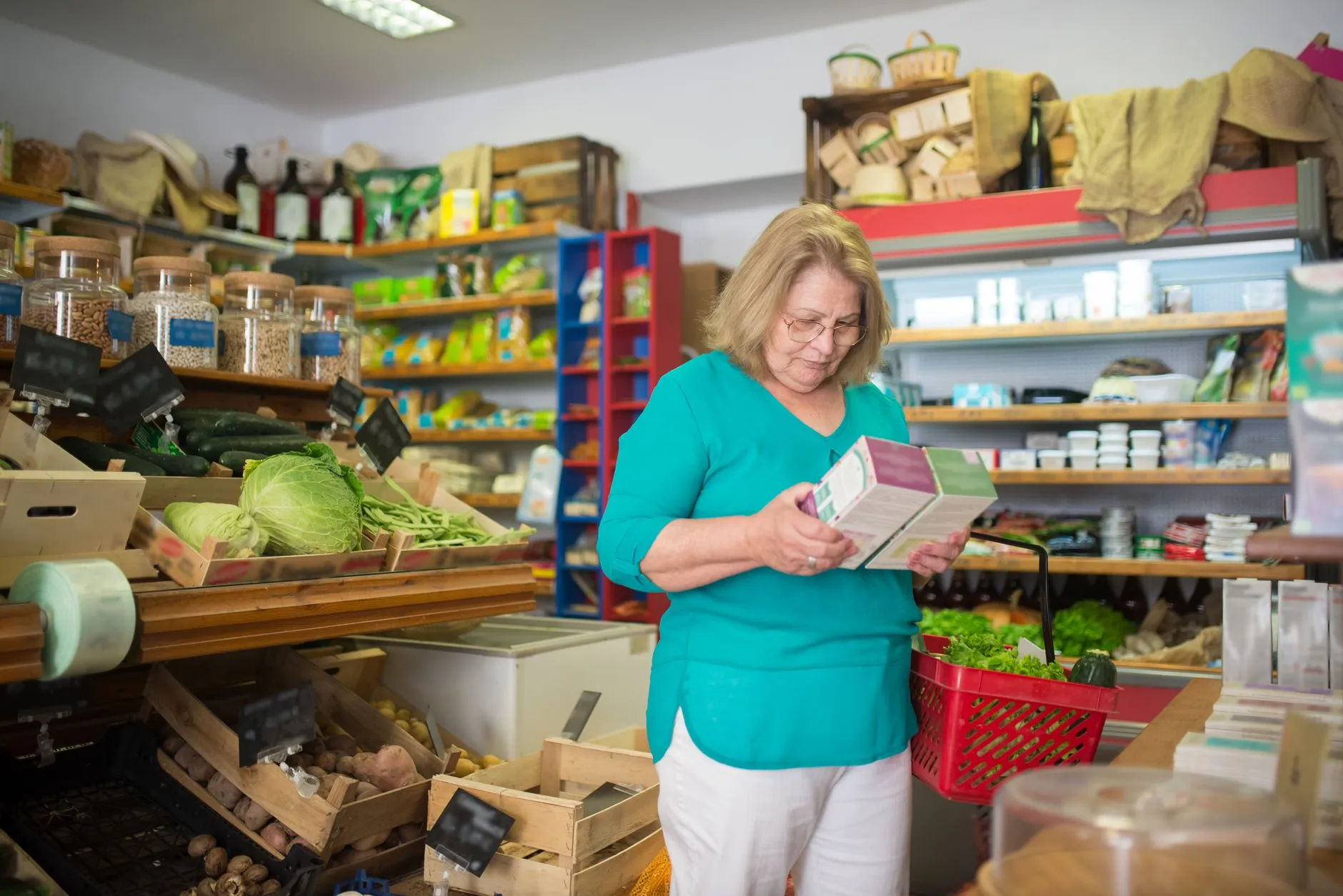 An older woman in a teal top compares two boxes of food in a small grocery store, a red basket on her arm.