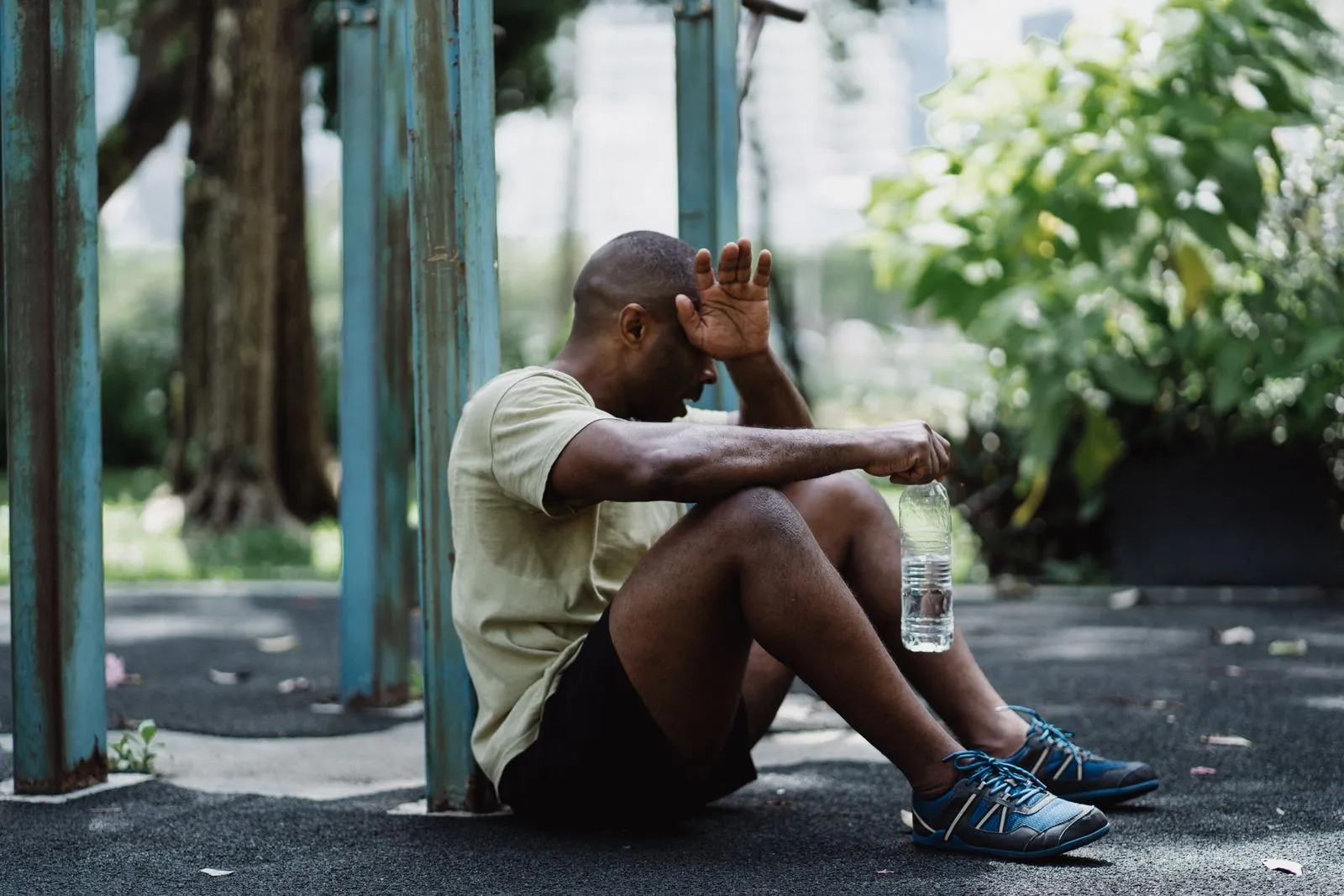 A man in a green shirt sits on the ground in a park, wiping his forehead with his forearm, a water bottle in one hand.