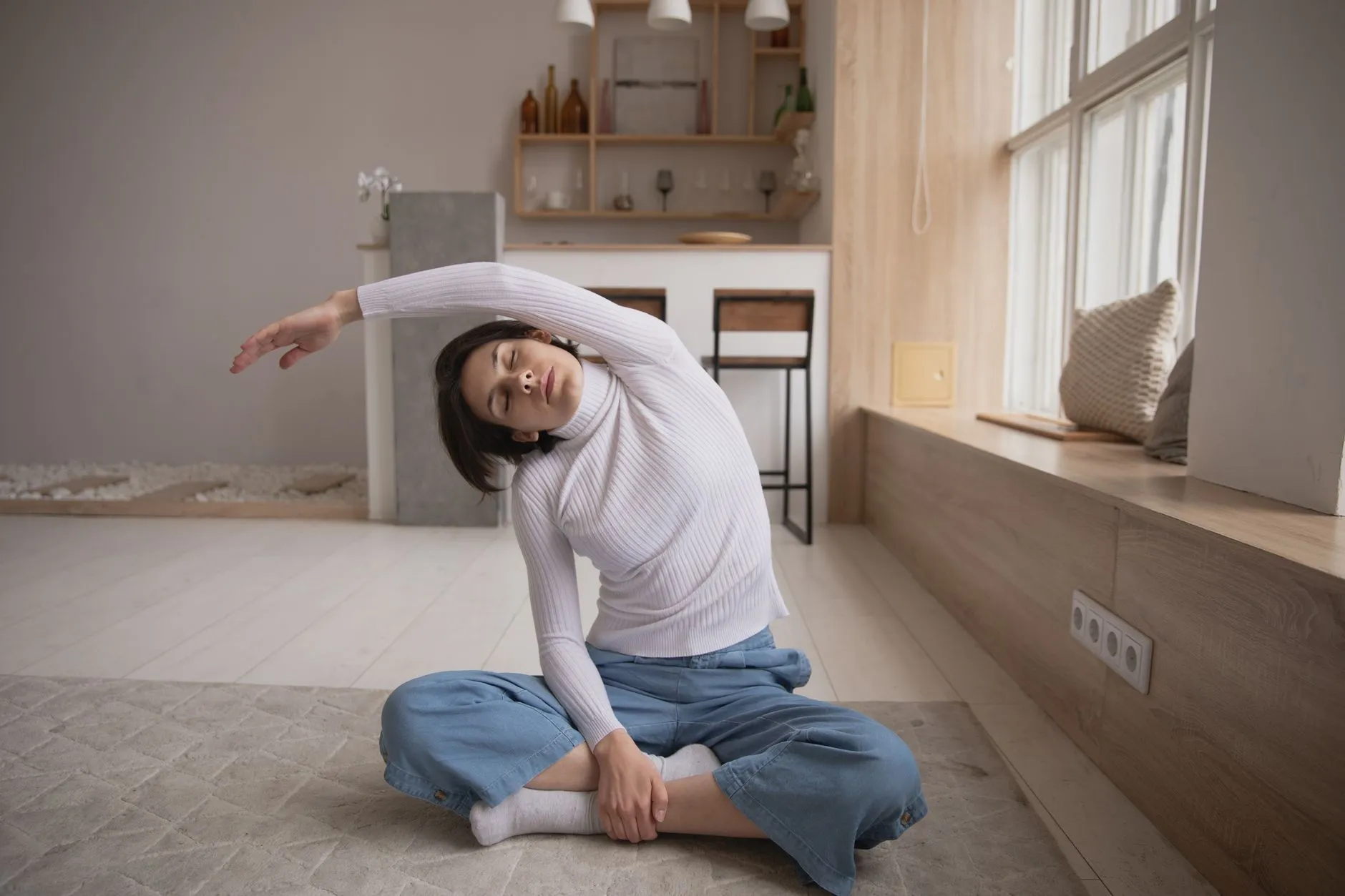 A woman in a sweater and jeans sitting on a rug at home, eyes closed, doing a relaxed side stretch in a bright living room.